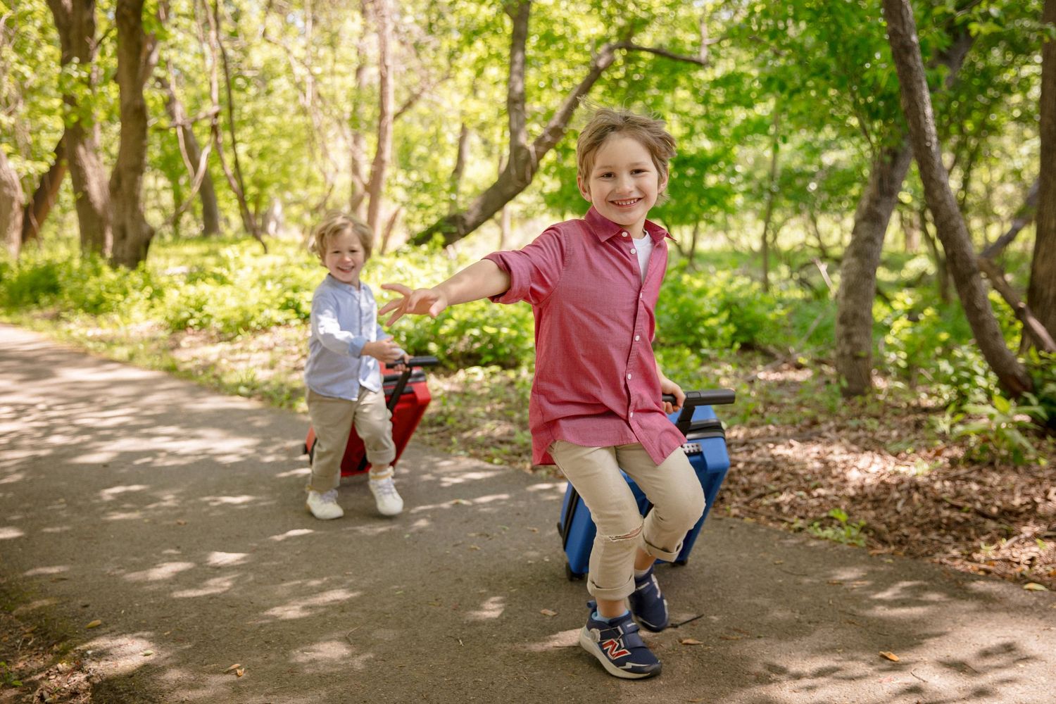 Two happy boys riding toy bikes on a sunny park path surrounded by lush green trees.