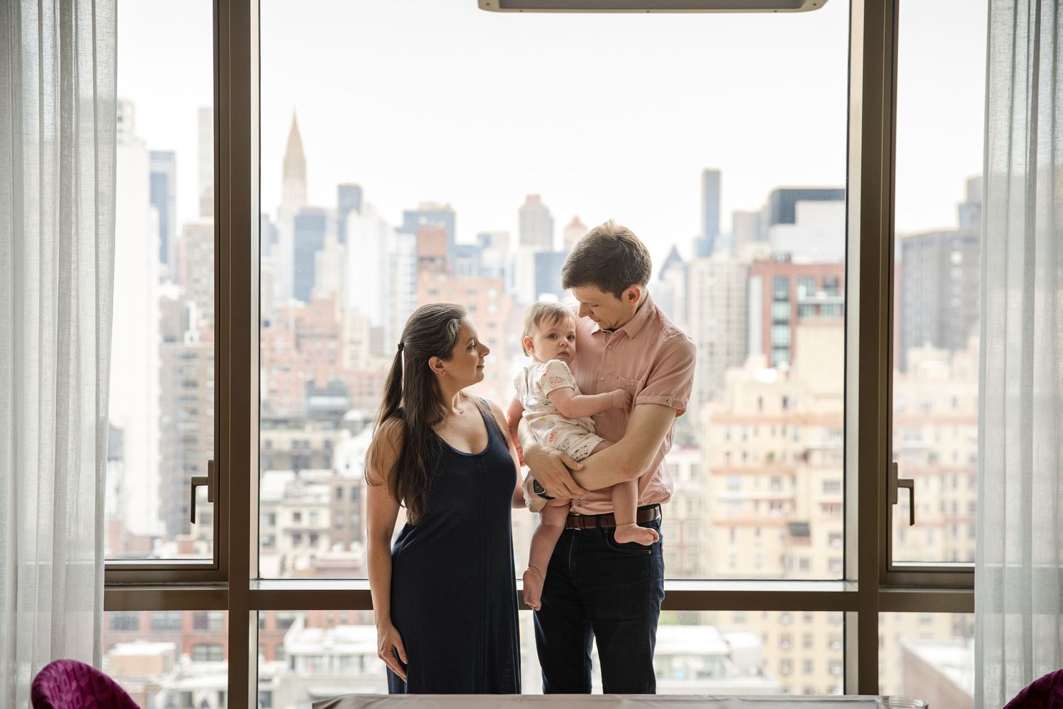 Young family with baby stands by large city view window in modern apartment with NYC skyline backdrop.