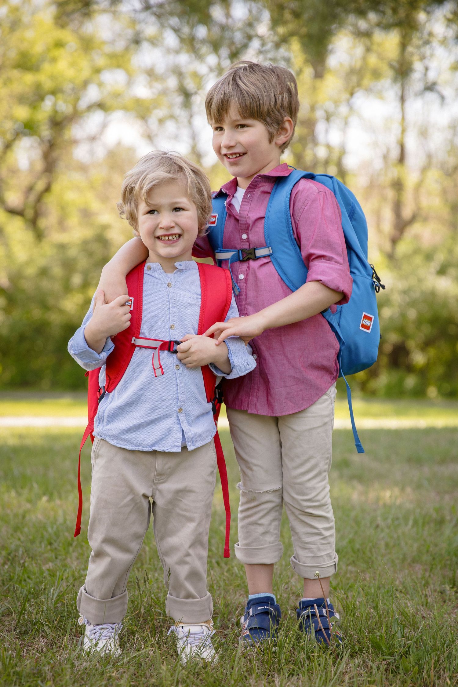 Two smiling boys with backpacks stand together outdoors, ready for school on a sunny day.