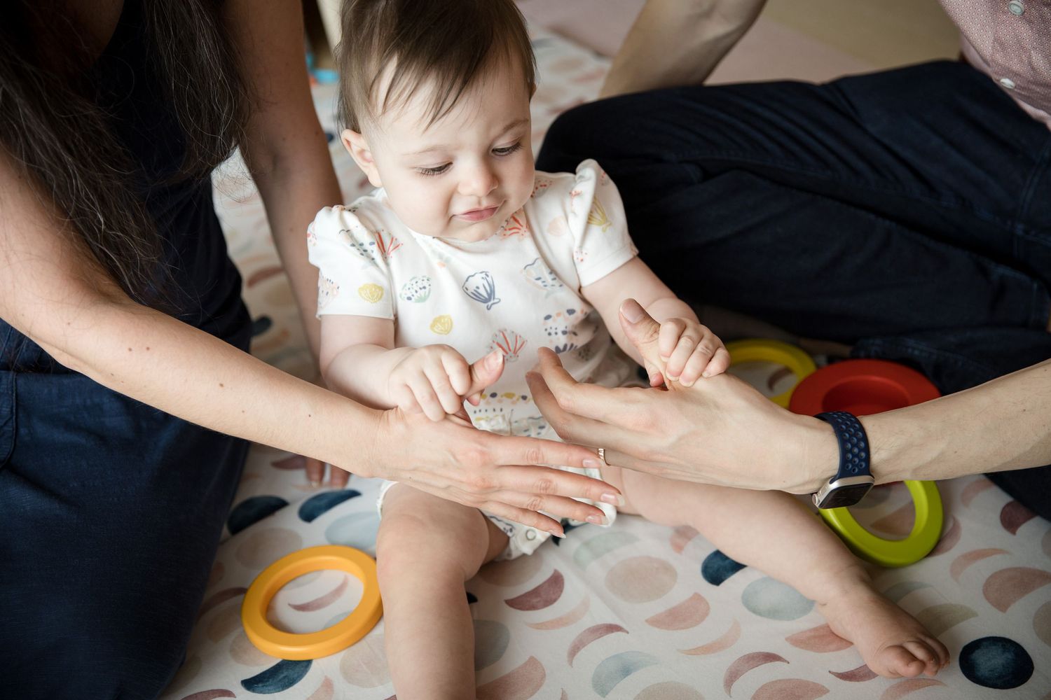 Baby sitting on a play mat with two adults gently holding their hands during a developmental play session.