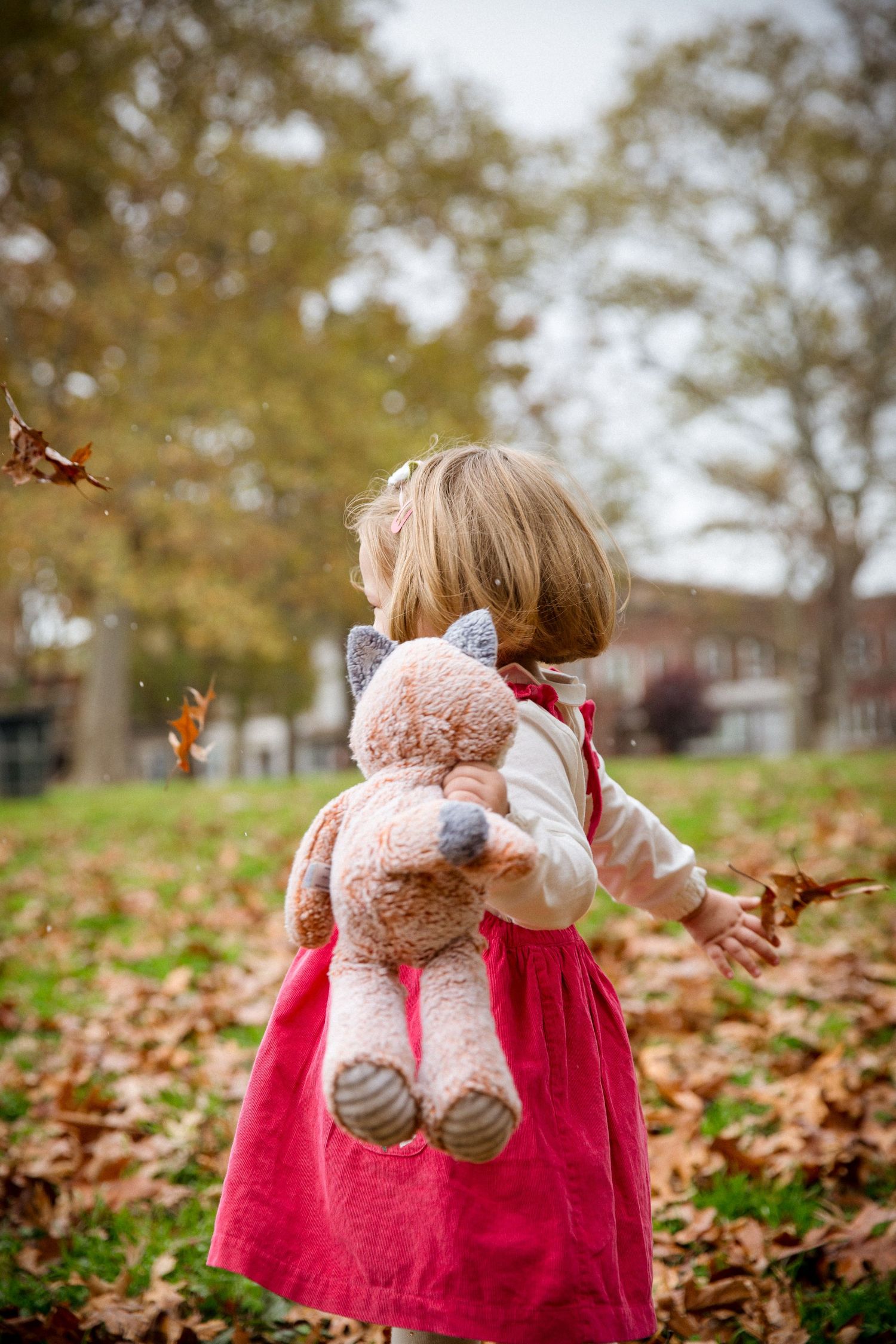 Toddler girl in pink dress holding stuffed unicorn toy while playing in autumn leaves at park.