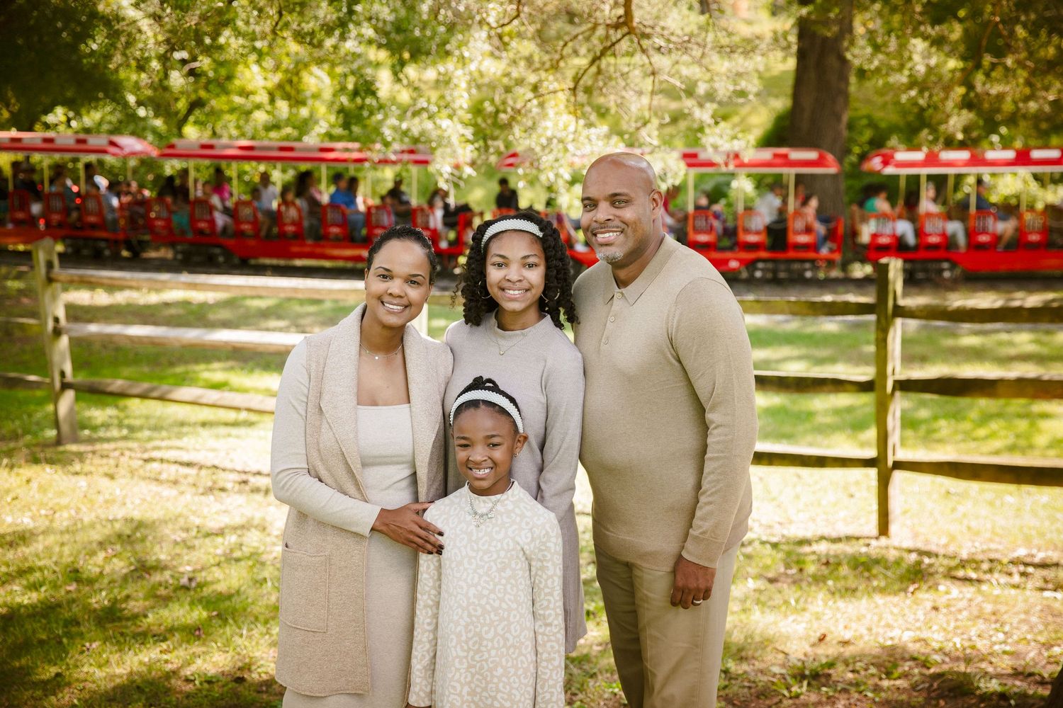 Smiling family of four poses together in a sunny green park near a red train ride.
