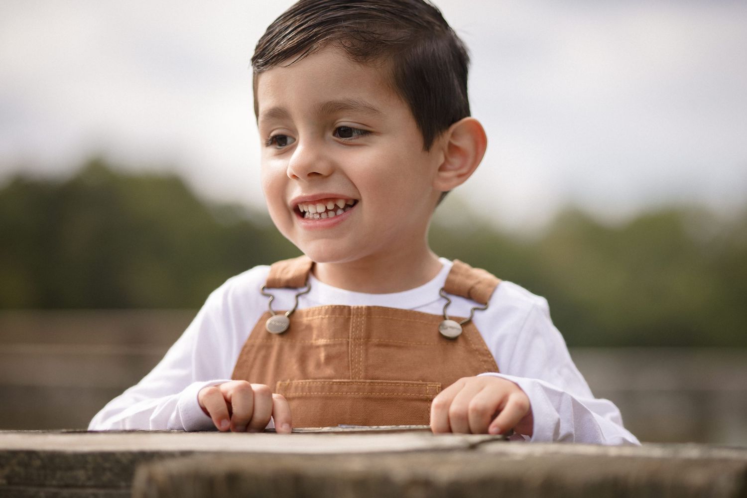 Smiling young boy in brown overalls and white shirt leaning on a ledge outdoors.