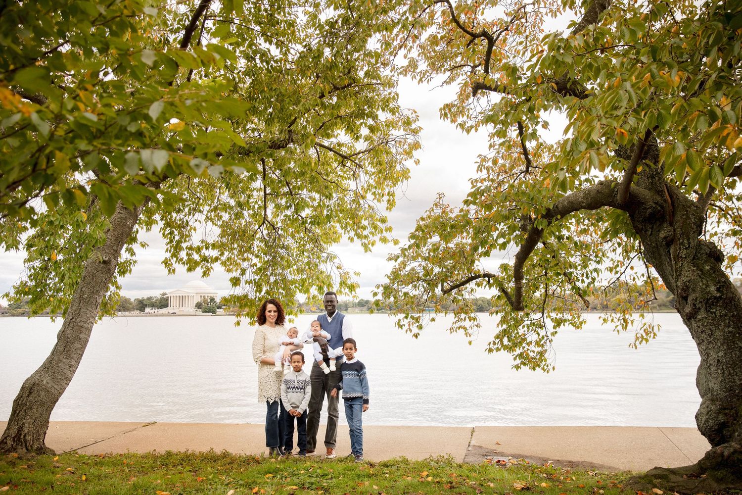 Family of six stands beneath large trees near a waterfront with a monument in the background.