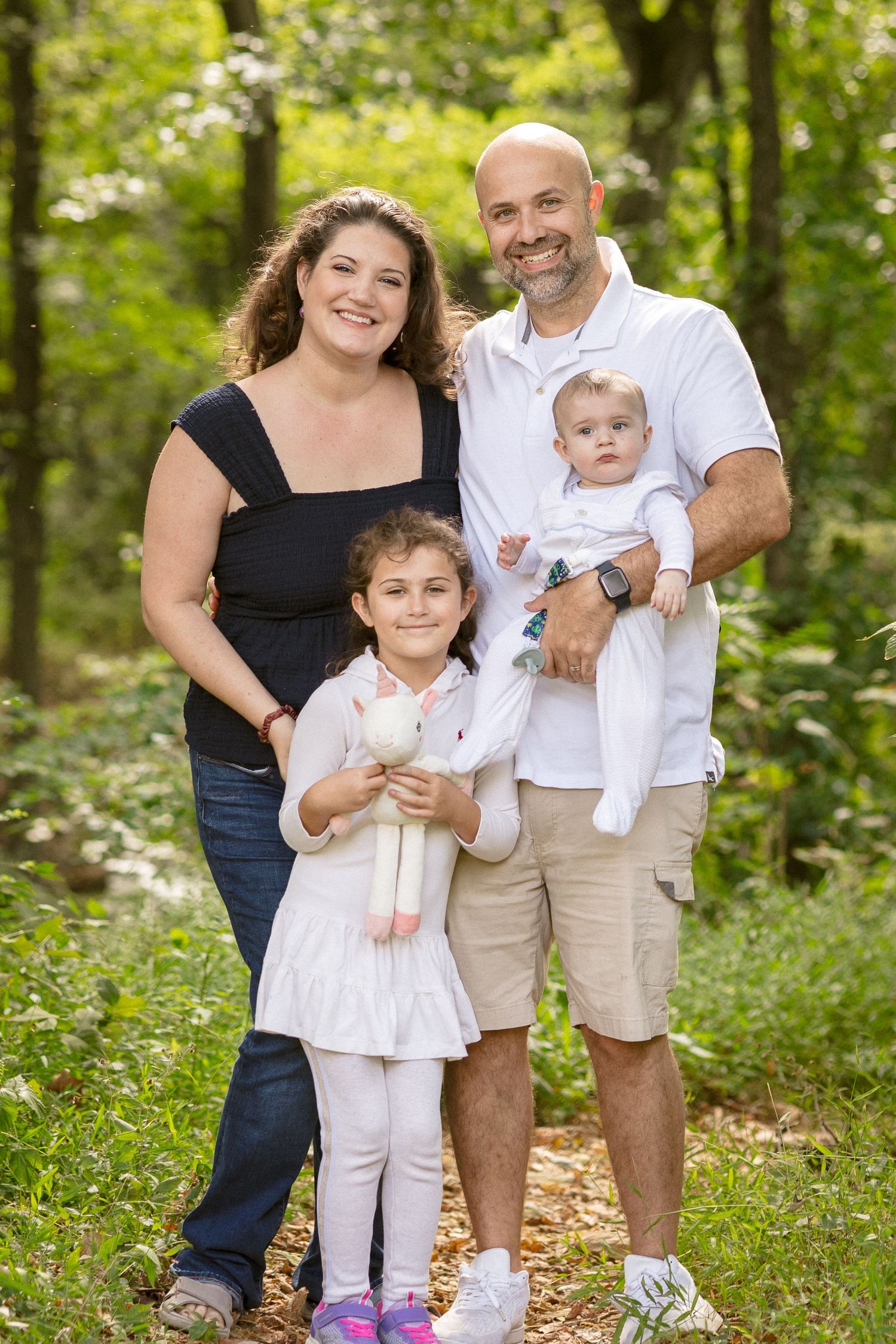 Happy family of four posing outdoors in lush green forest, parents smiling with young daughter holding stuffed bunny and baby.