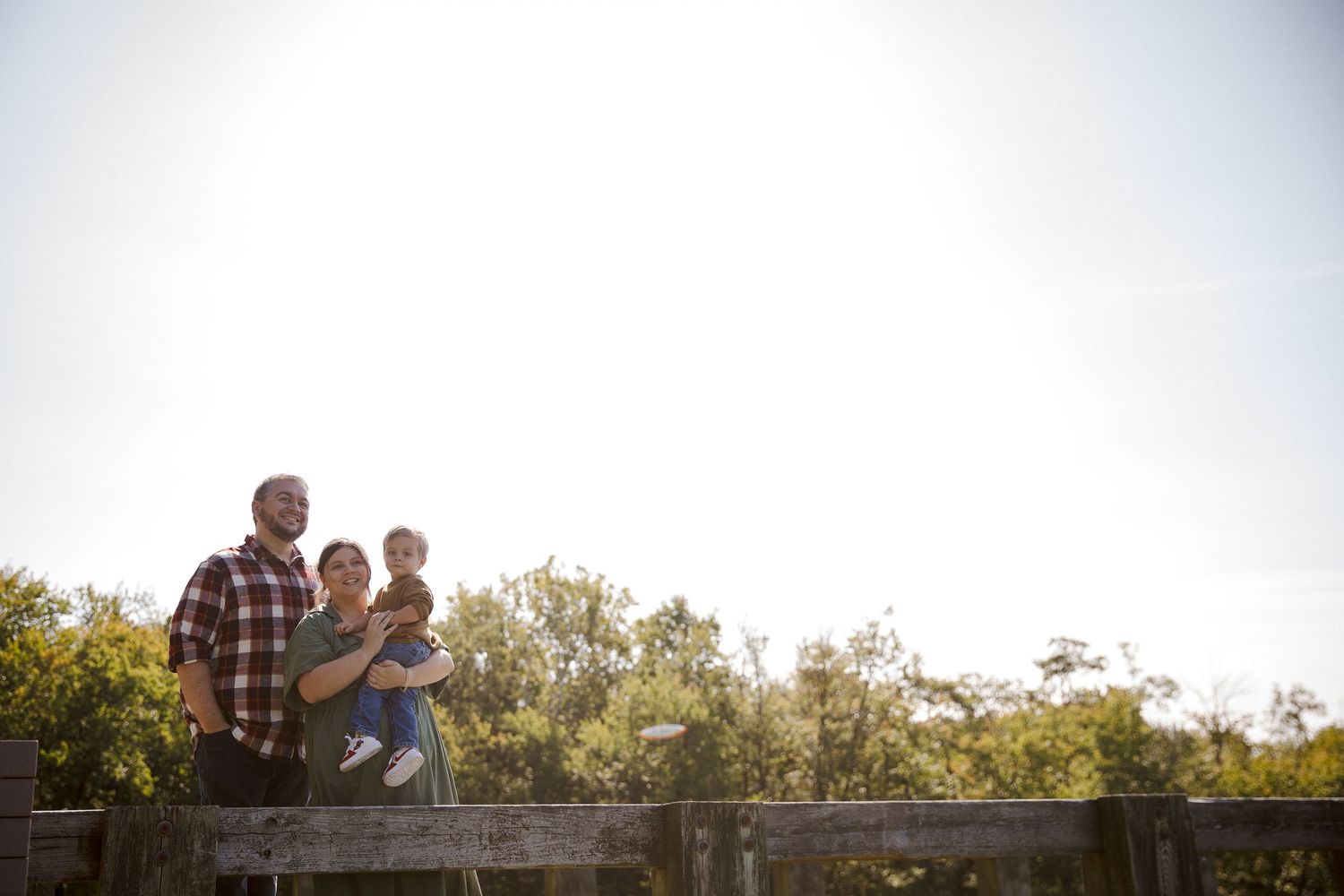Family of three stands on wooden bridge overlooking green treetops against bright sky.