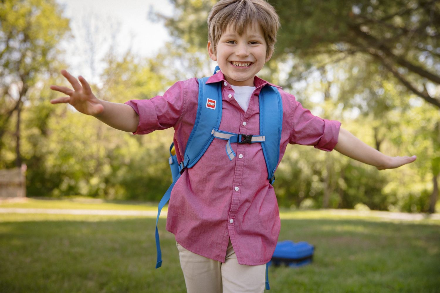 Happy young child wearing a pink shirt and blue backpack smiling with arms outstretched in a green park.