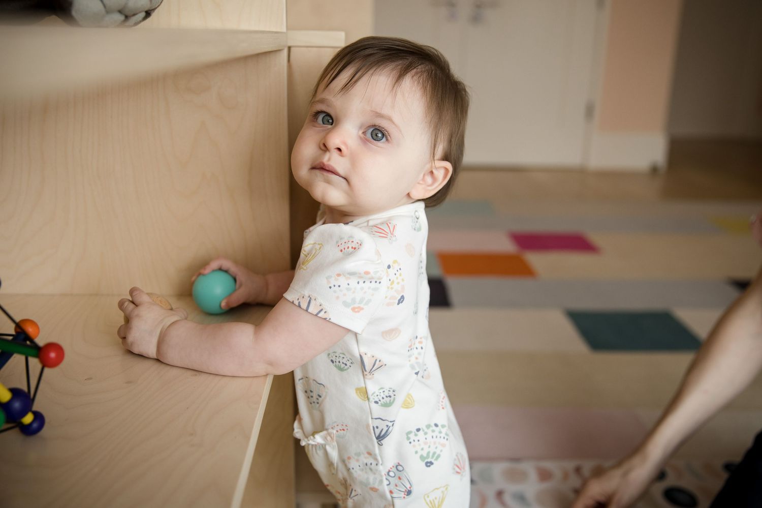 Curious baby standing and holding onto a wooden furniture piece, wearing a white onesie in a colorful playroom.