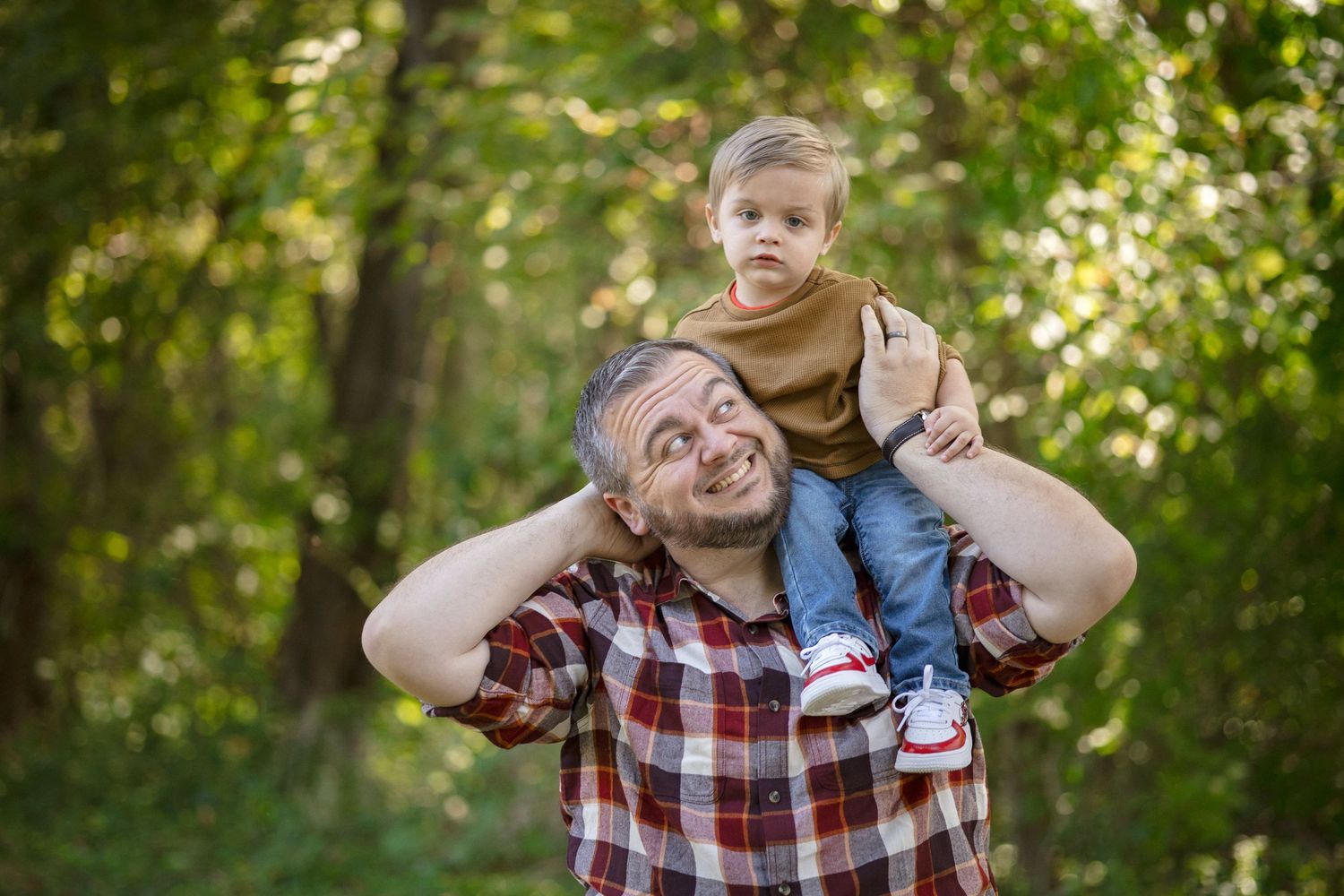 A smiling father carries his young toddler son on his shoulders in a lush green forest setting.