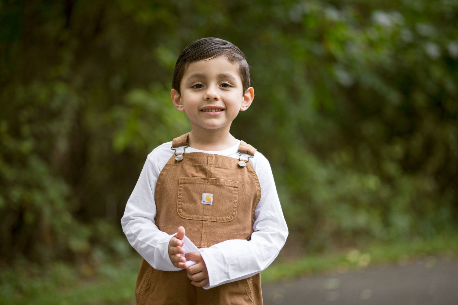 Young boy in brown Carhartt overalls and white shirt smiling outdoors with green trees in background.