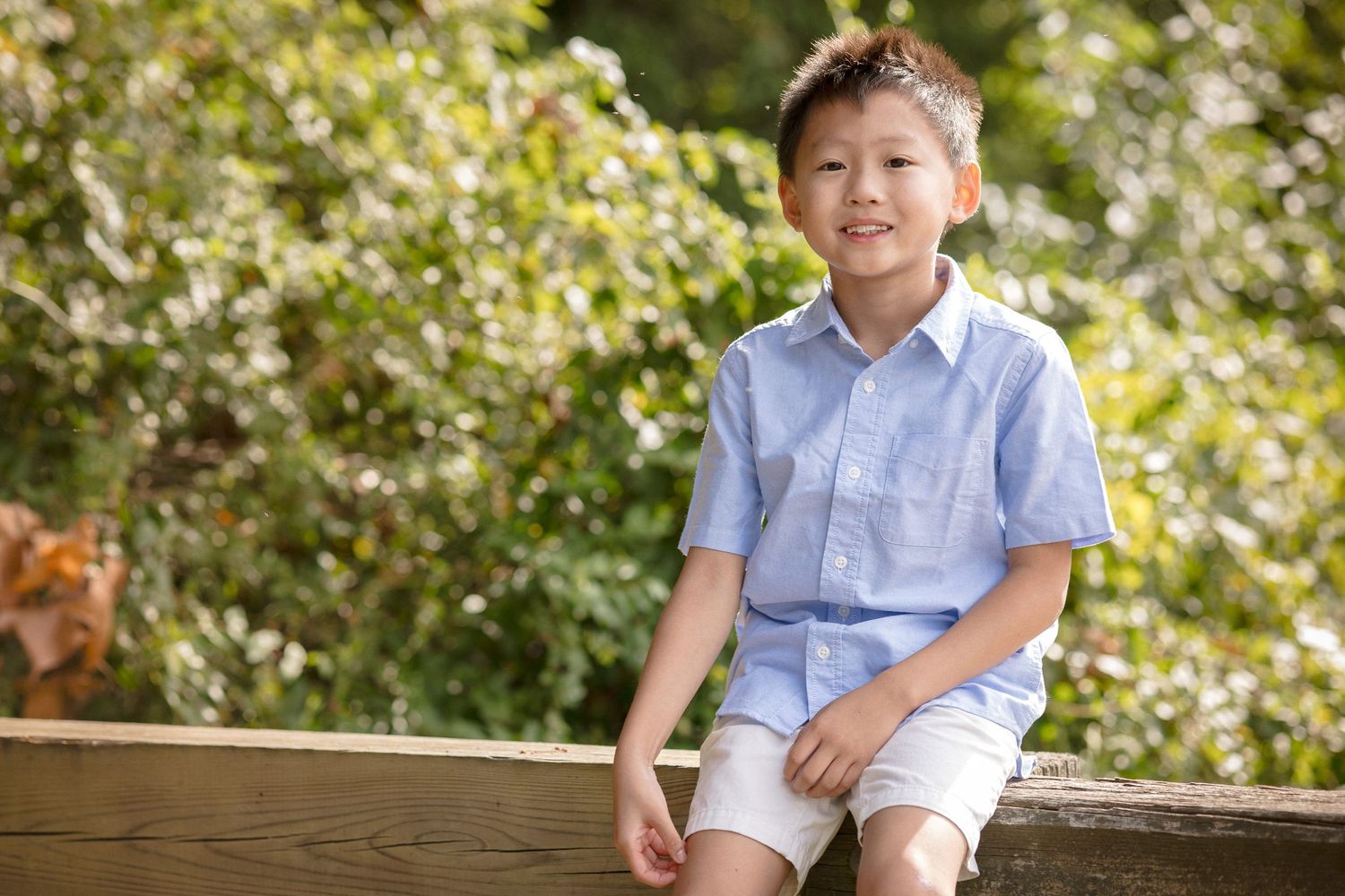 A young Asian boy in a light blue shirt and white shorts sits smiling outdoors with green trees behind him.