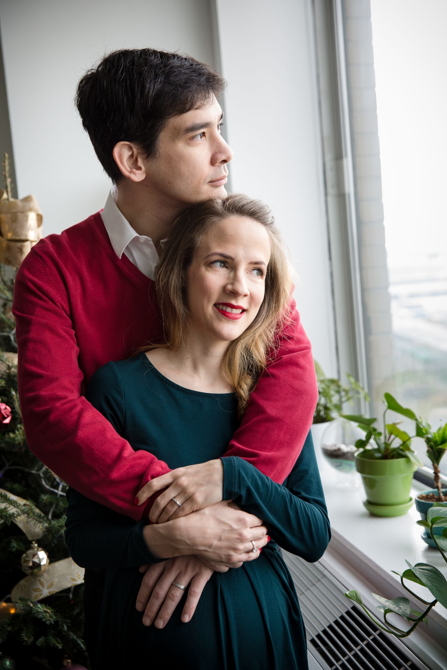 Couple embracing by window near Christmas tree, man in red sweater hugging woman in teal dress.