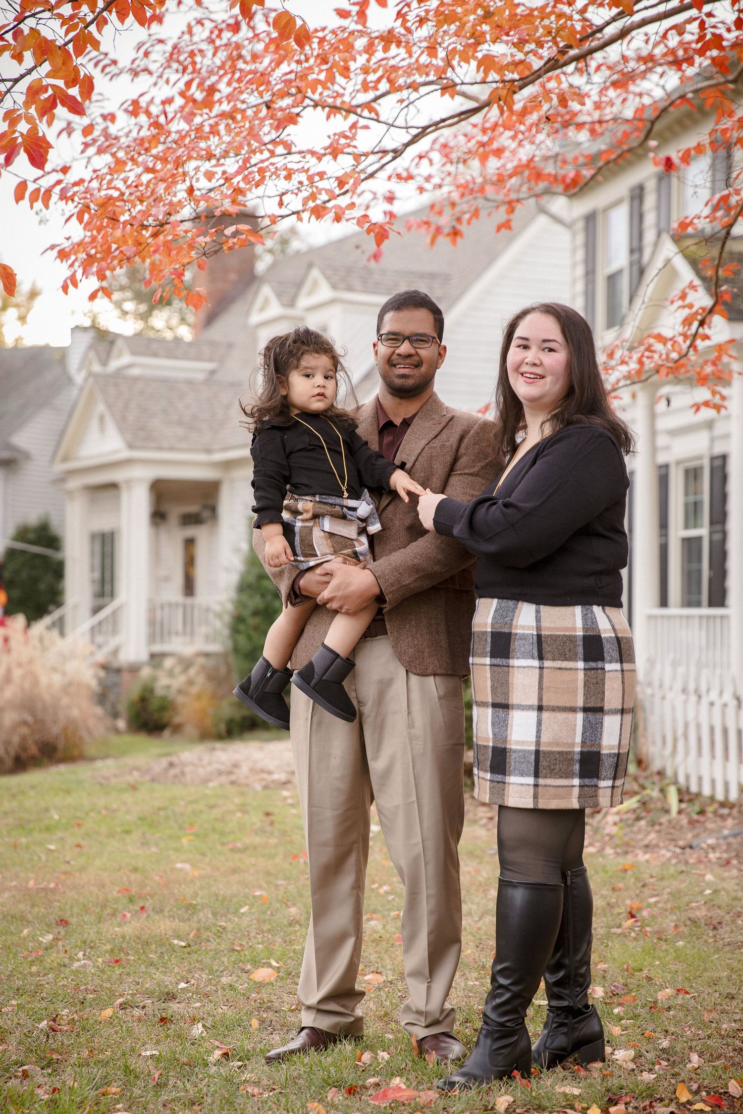 Family of three posing outdoors in autumn, man holding toddler beside woman, white house and red foliage in background.