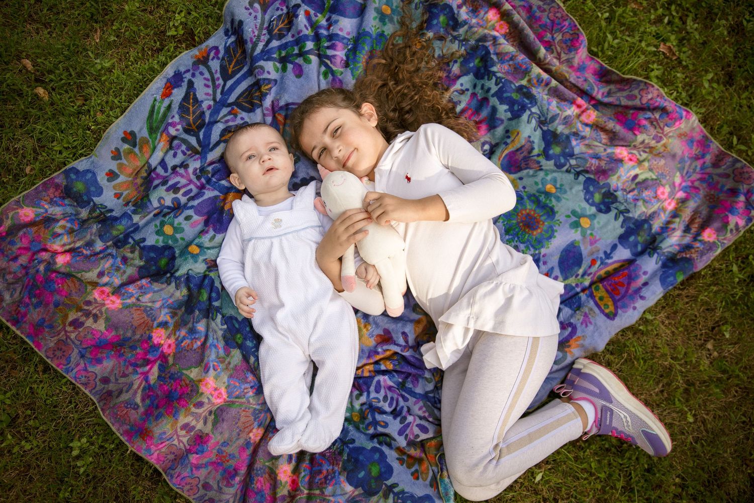 Two sisters lying on a colorful floral blanket outdoors, older girl hugging younger baby sibling.