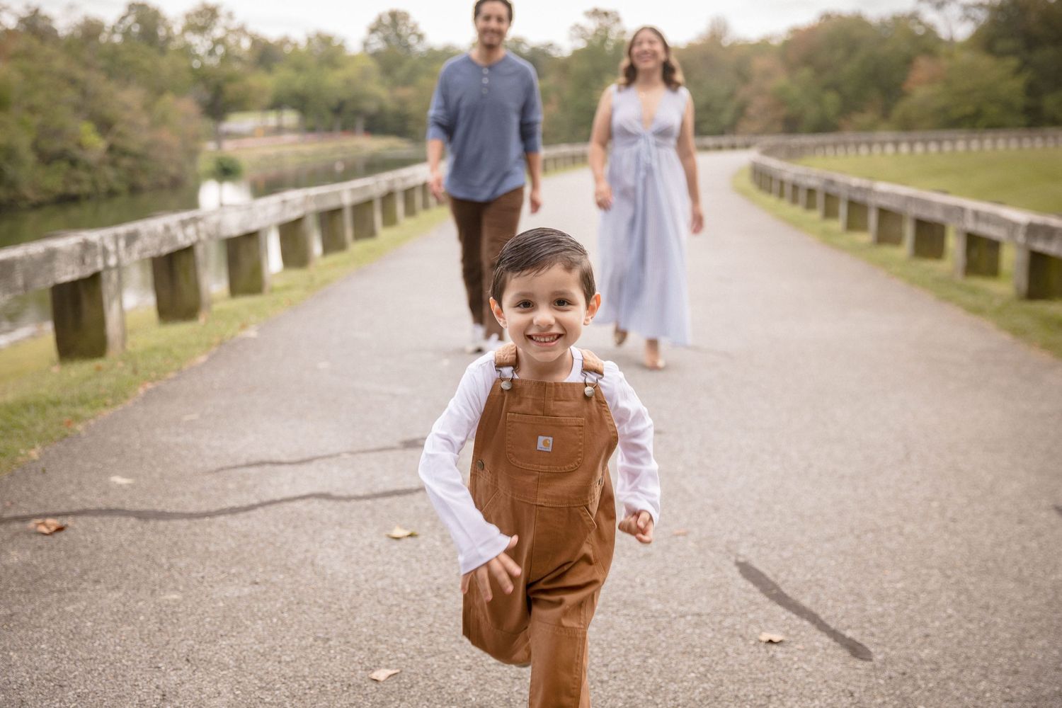 A joyful young boy in brown overalls runs toward the camera as his parents walk behind him on a tree-lined path.