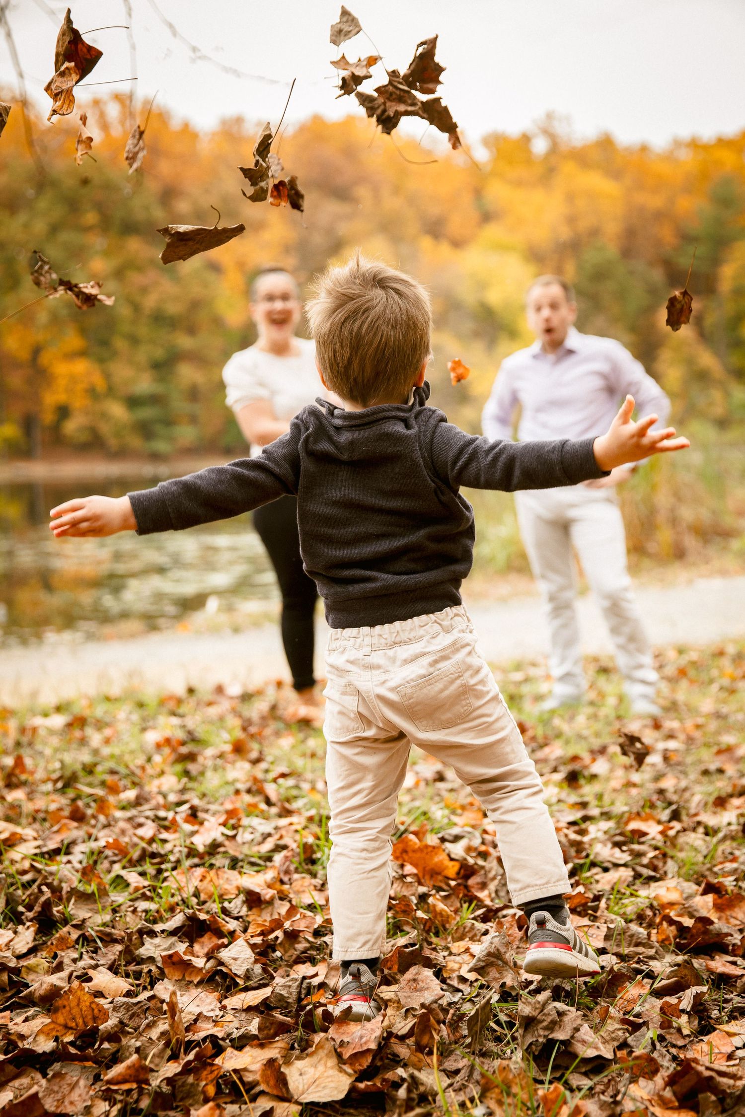Excited toddler tosses autumn leaves while parents watch joyfully in a park.