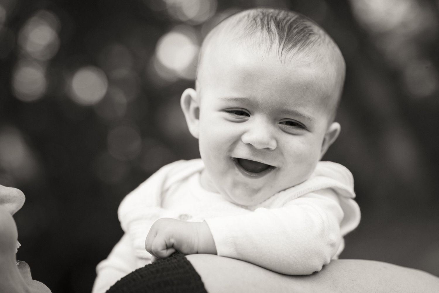 Joyful baby smiling brightly in black and white portrait photo, held up by adult hands outdoors.