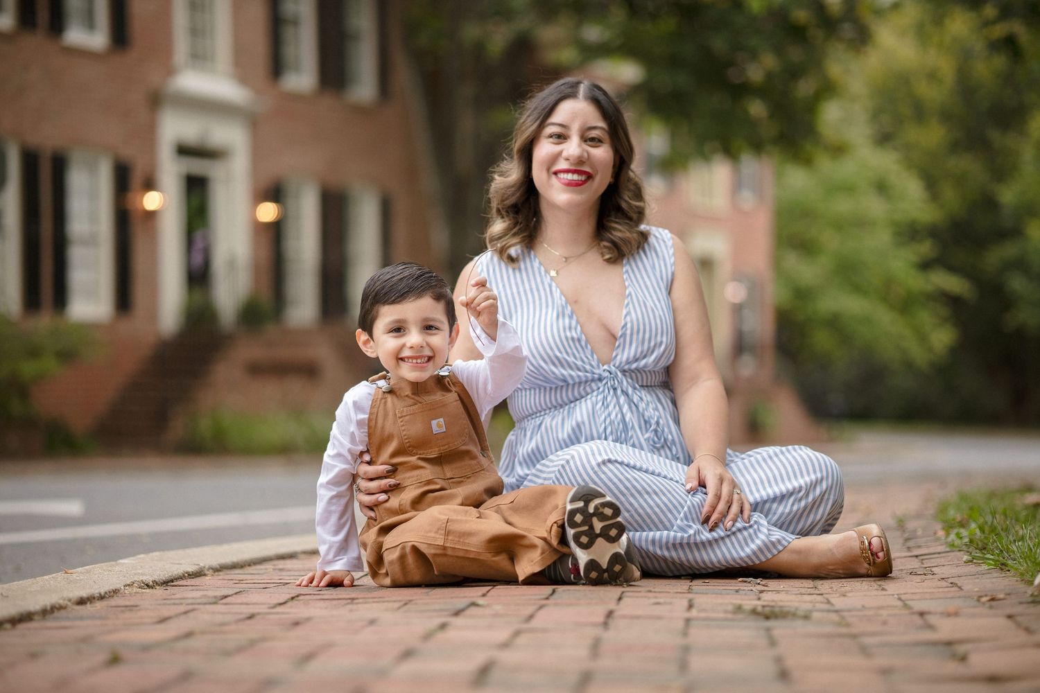 A woman and young boy sit smiling on a brick path outdoors, she wears a striped jumpsuit, he wears brown overalls.