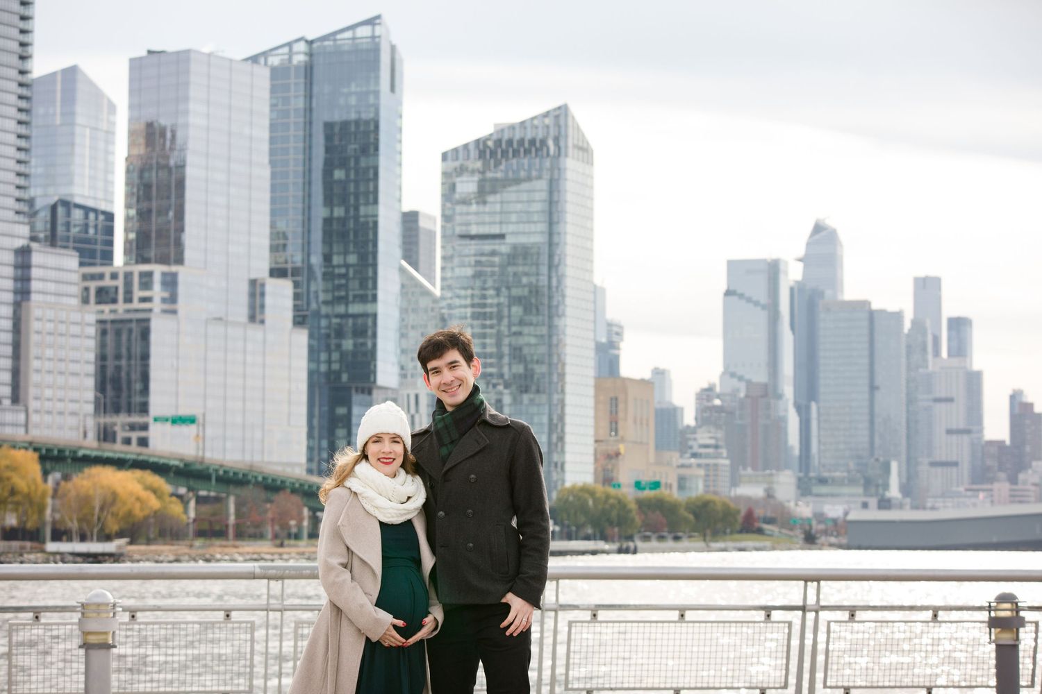 Couple posing together on NYC waterfront with Manhattan skyline skyscrapers in background on cloudy day.