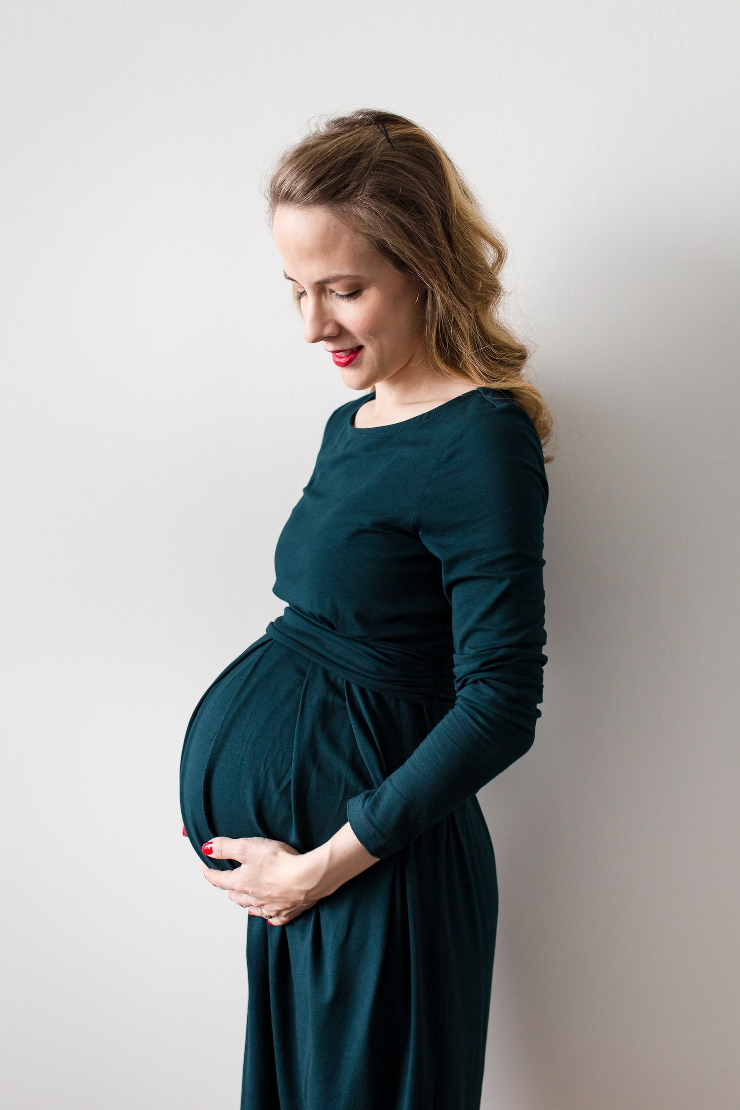 Pregnant woman in green dress cradling baby bump, smiling downward against white background.