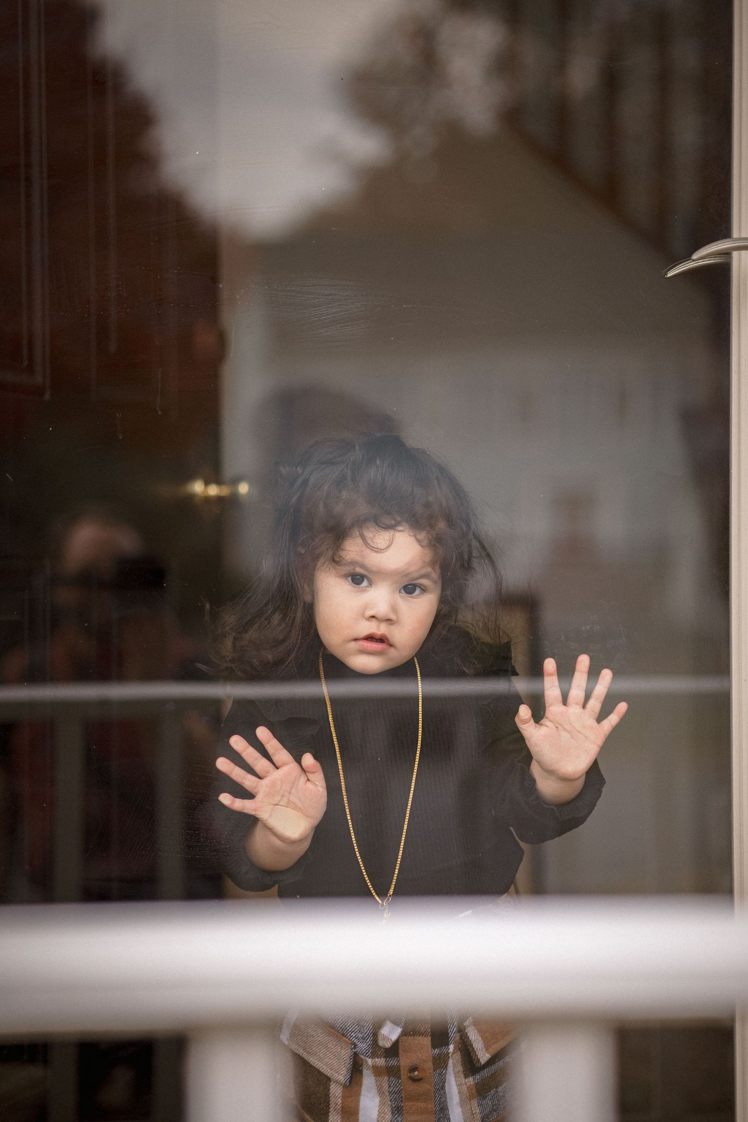 Young child with curly hair and gold necklace pressing hands against glass door, looking outside with wide eyes.