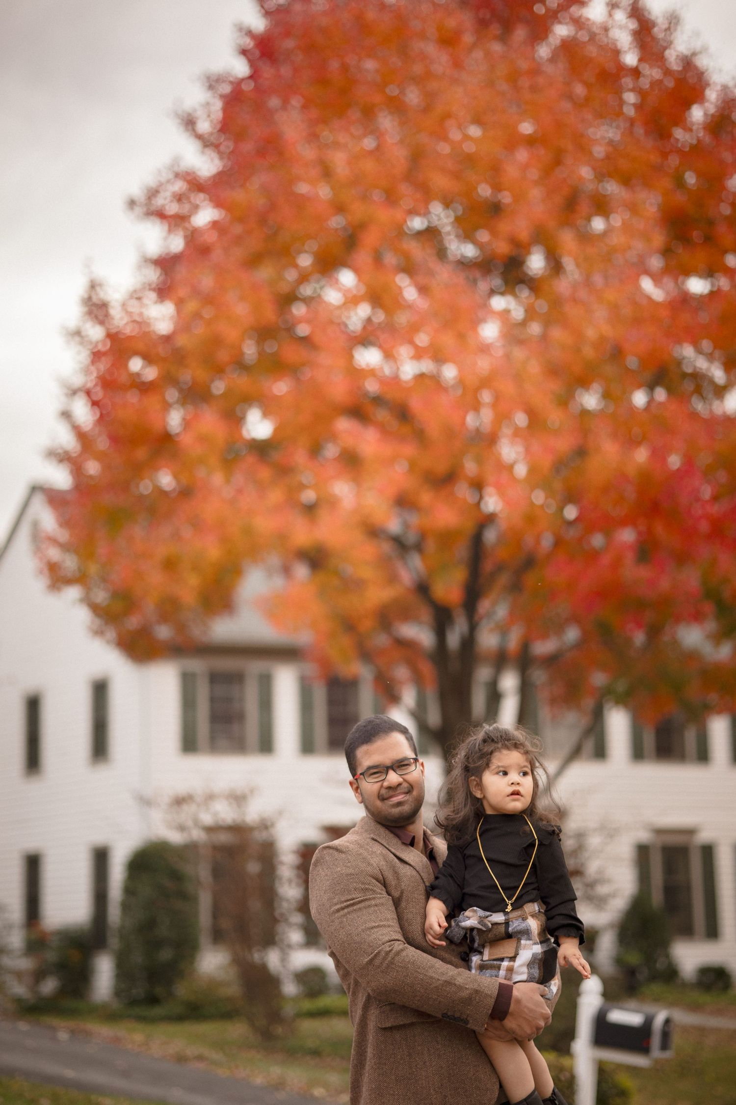 Father holding young child outdoors in autumn, vibrant red maple tree and white house in background.