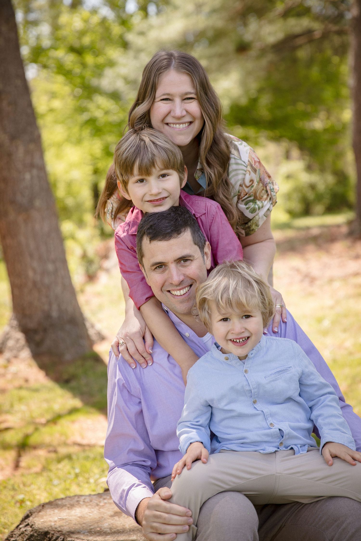 Happy family of four posing outdoors in a sunny park, with parents and two young boys smiling together.