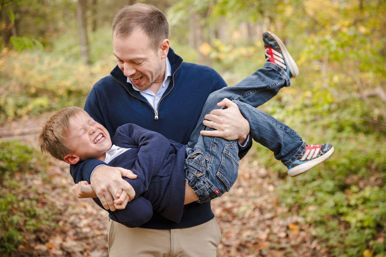 Laughing father playfully carries his young son horizontally through an autumn forest path.