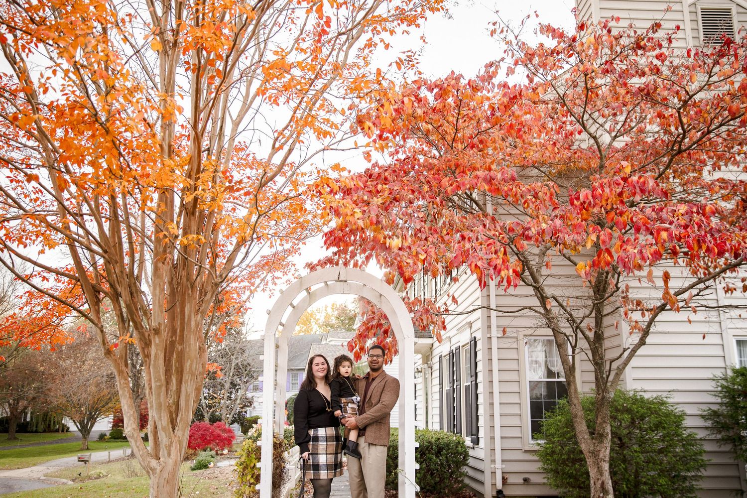 Family of three poses under a white garden arch surrounded by vibrant red autumn trees.