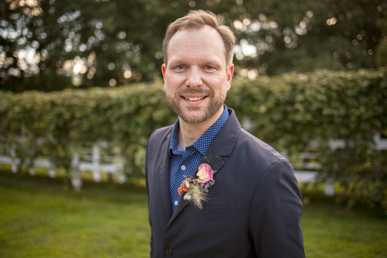 Smiling man in navy blazer wearing a floral boutonniere outdoors at an event.