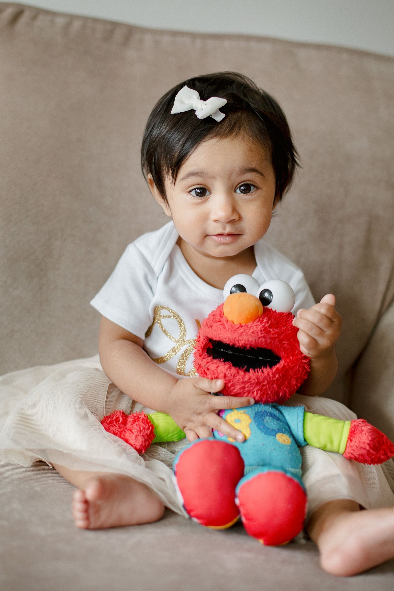 Cute baby girl holding an Elmo stuffed toy while sitting in a beige armchair.