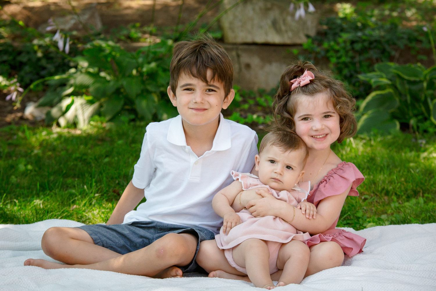 Three siblings posing together on a white blanket in a sunny garden, oldest holding the baby.