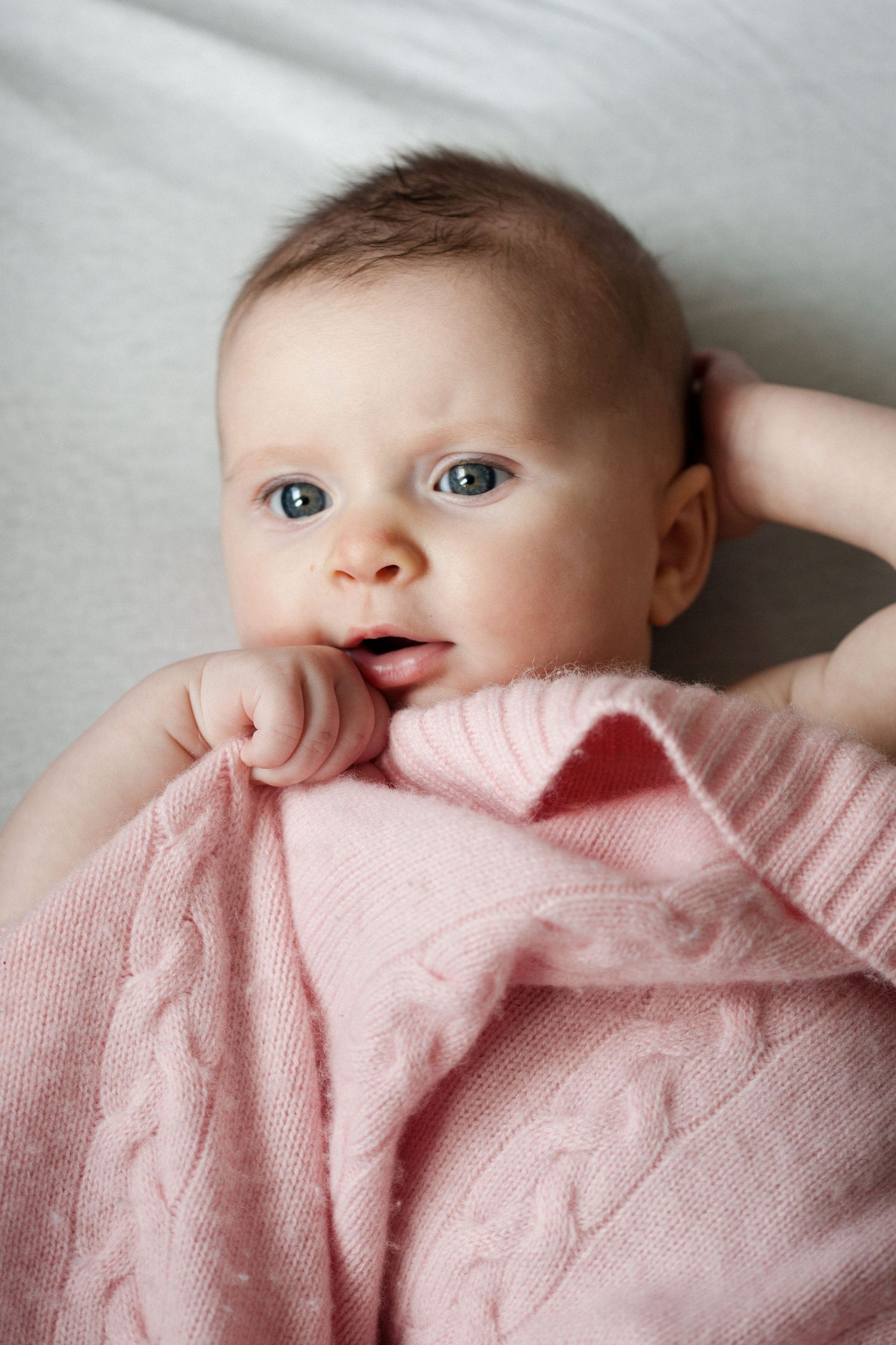 Adorable blue-eyed baby wrapped in a pink knit blanket lying on a white surface.