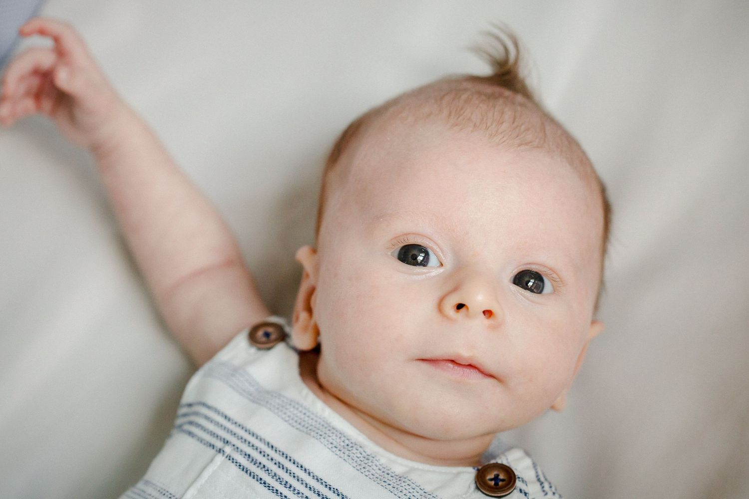 Newborn baby boy lying on white blanket with arm raised, looking up with curious wide eyes.