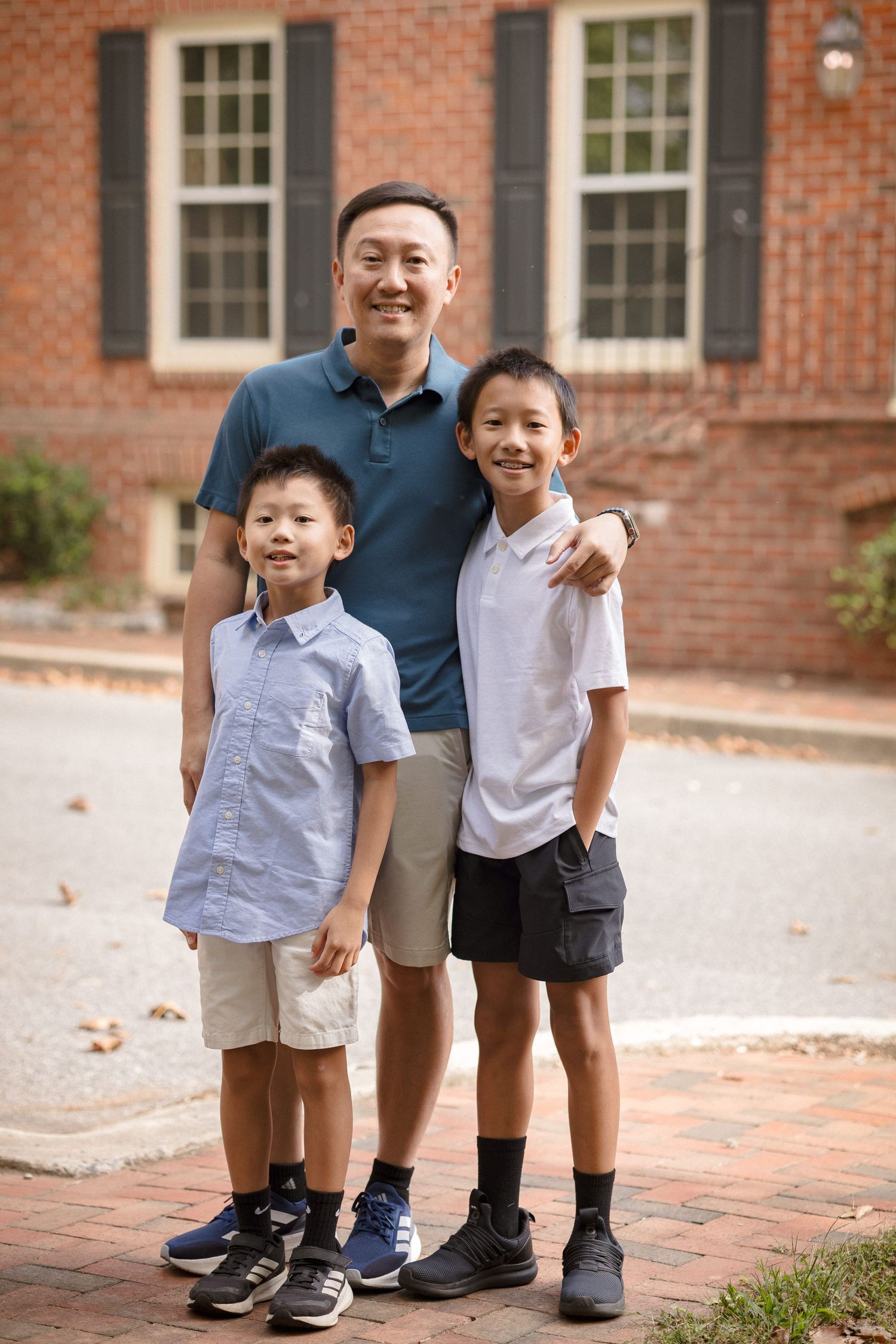 Asian father smiling with his two young sons outdoors in front of a brick building on a sunny day.