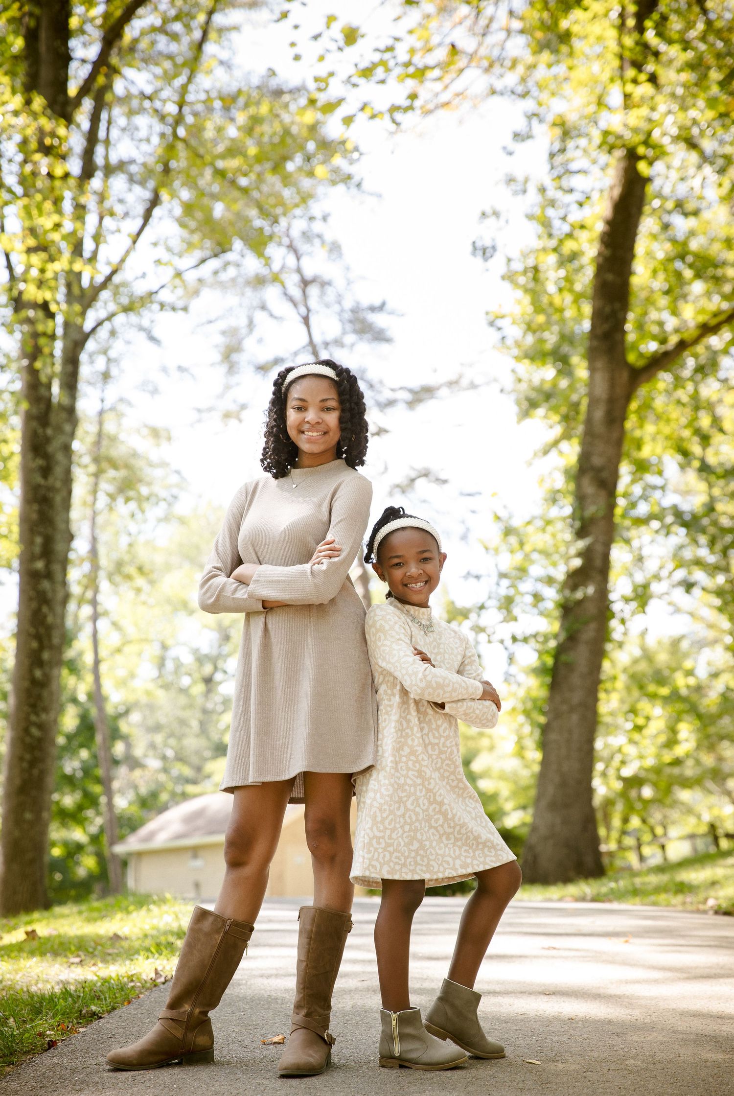 Two girls in matching beige dresses stand with arms crossed on a sunny tree-lined path.