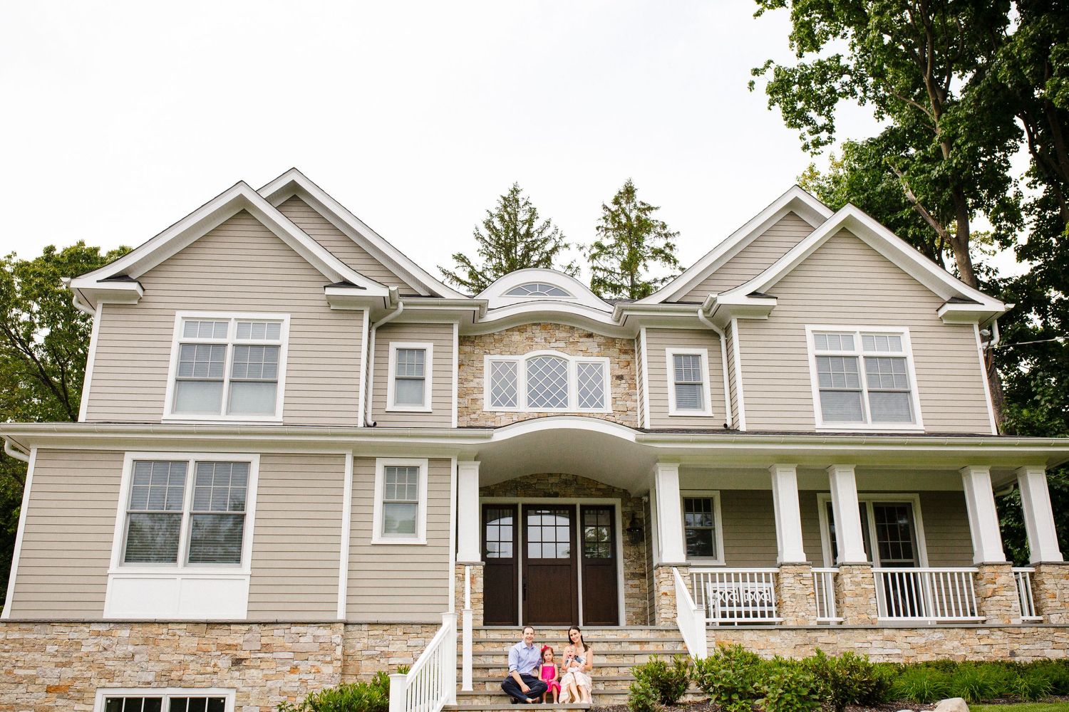 Large two-story suburban family home with stone accents, covered porch, and lush green trees.
