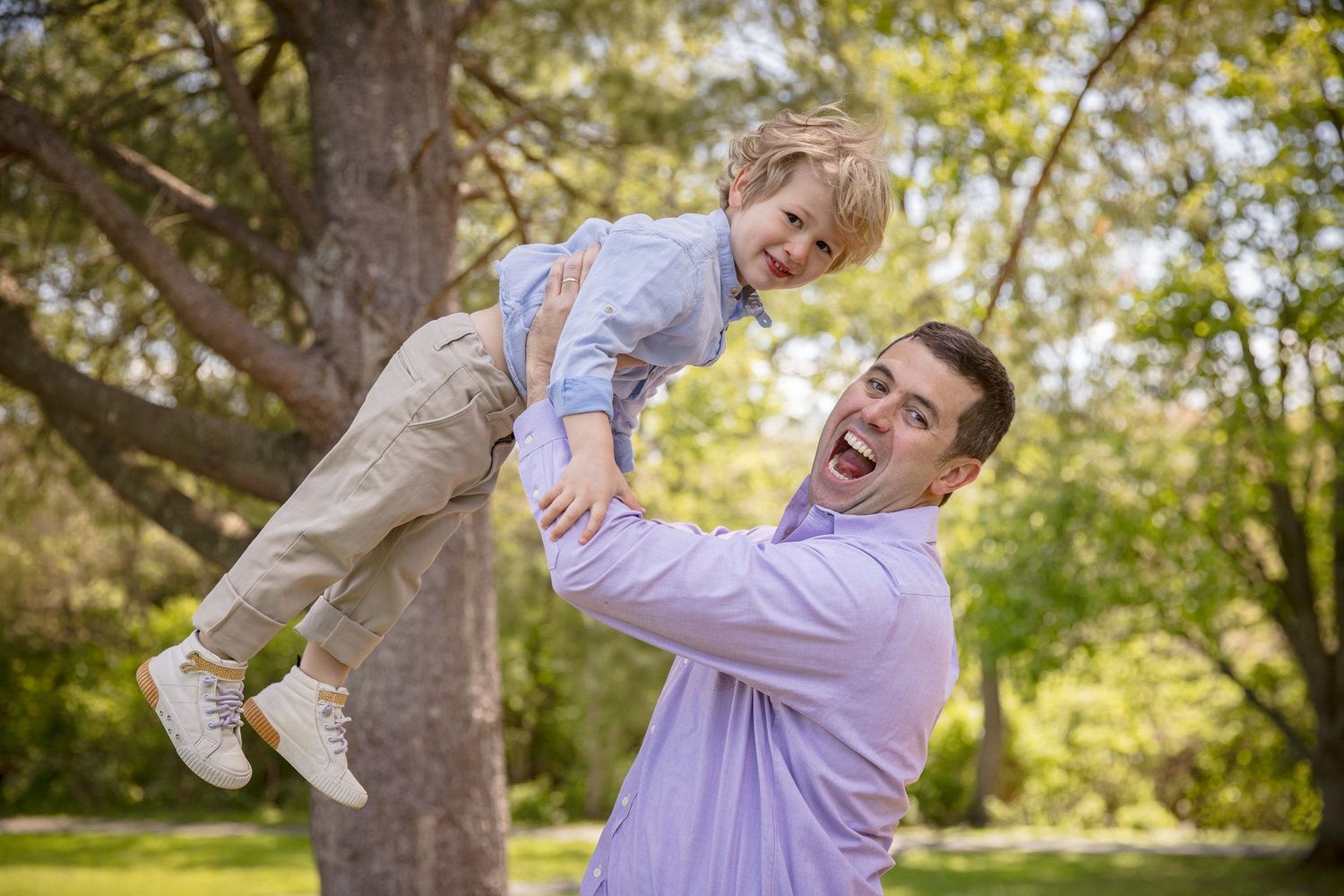 A joyful father lifts his laughing young son in a sunny green park, both smiling during outdoor family playtime.