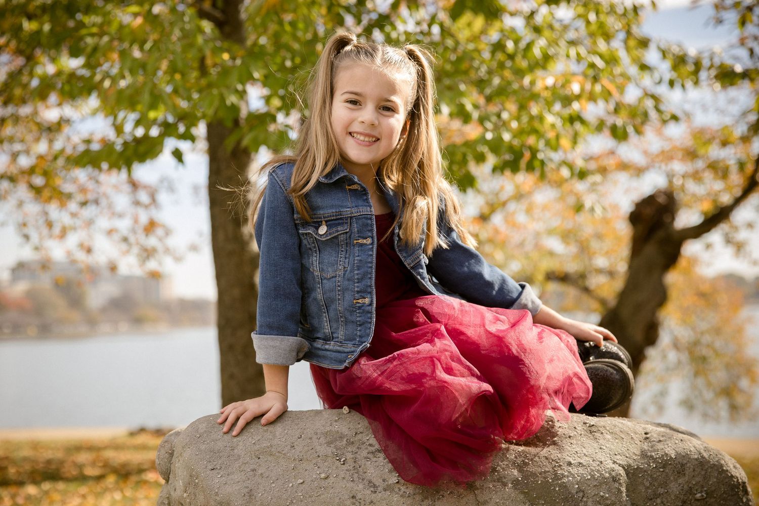 Blonde girl in denim jacket and red tulle skirt sitting on a rock in autumn park.