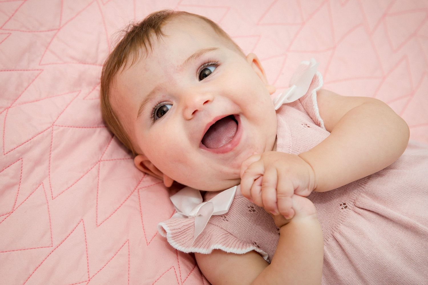 Joyful laughing baby girl in pink dress lying on pink blanket.