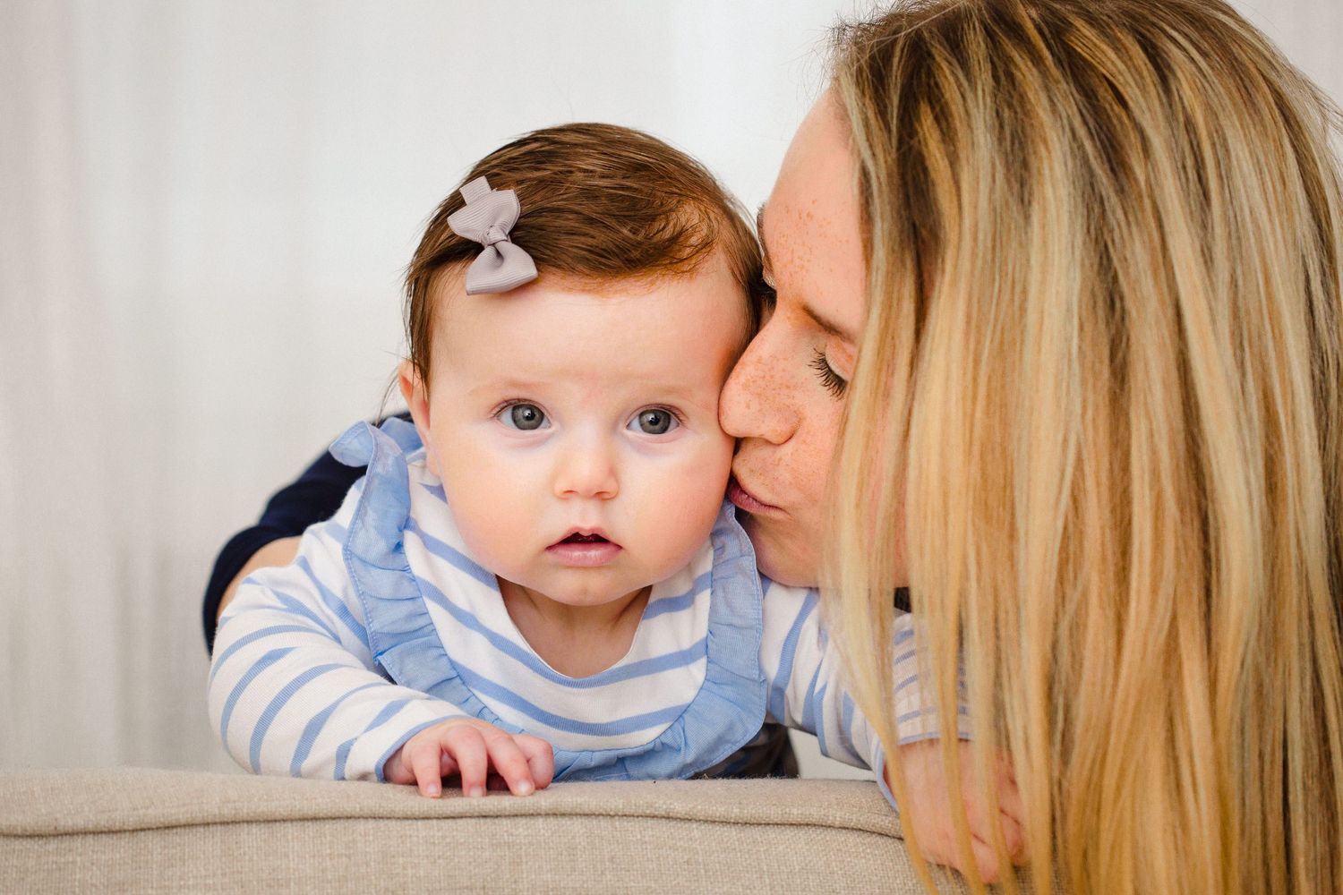 Blonde mother tenderly kissing her baby girl with bow in hair on cozy sofa.