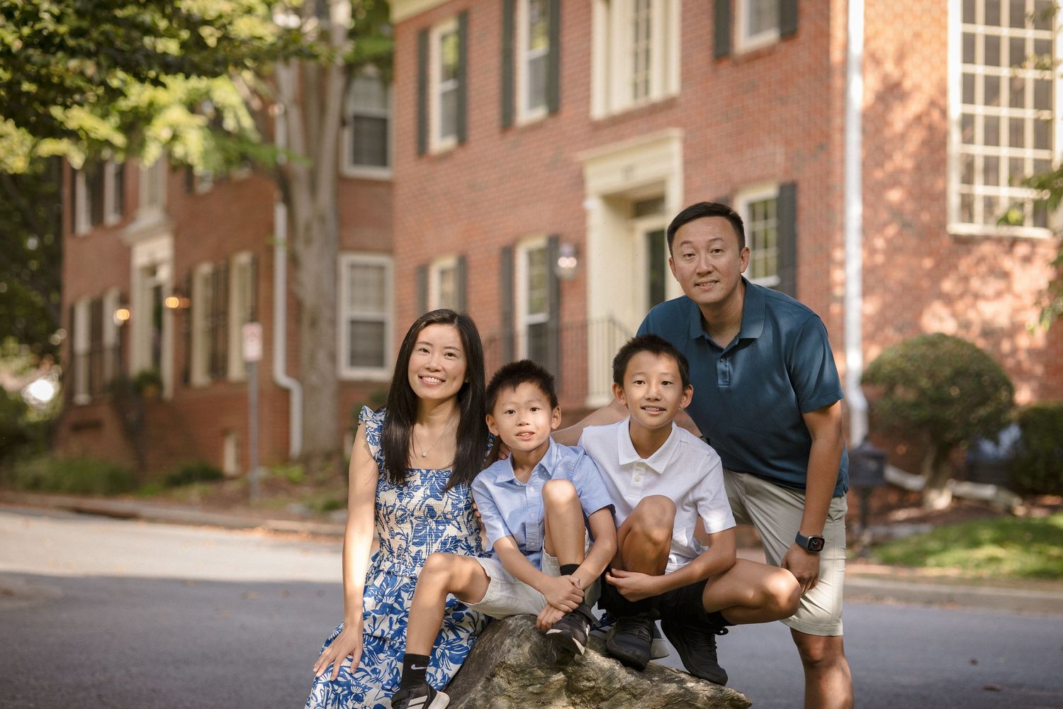 A smiling Asian family of four poses outdoors near a brick building, including two boys and their parents.