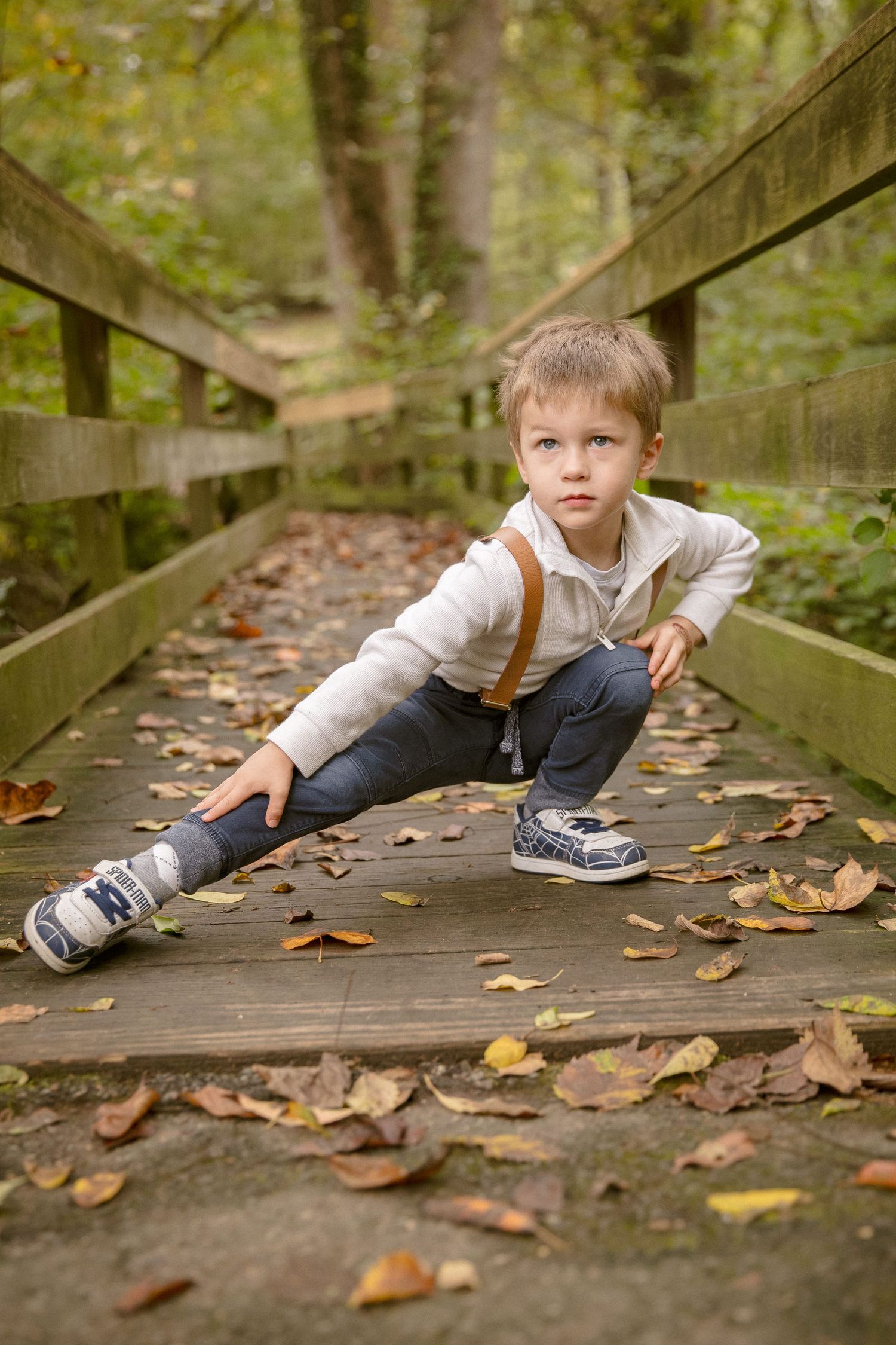 Young boy in suspenders crouching on a wooden bridge covered in autumn leaves in a forest.