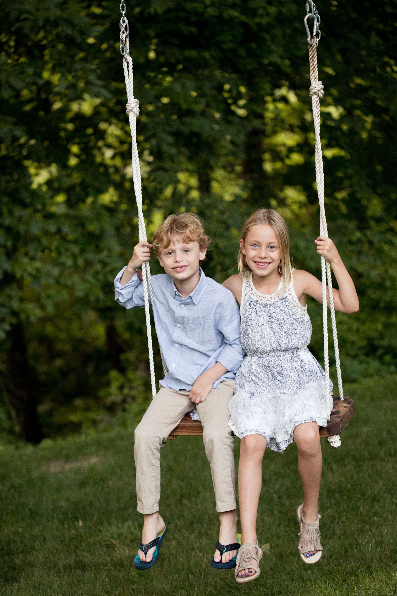 Two smiling children, a boy and girl, sitting together on a wooden rope swing outdoors.