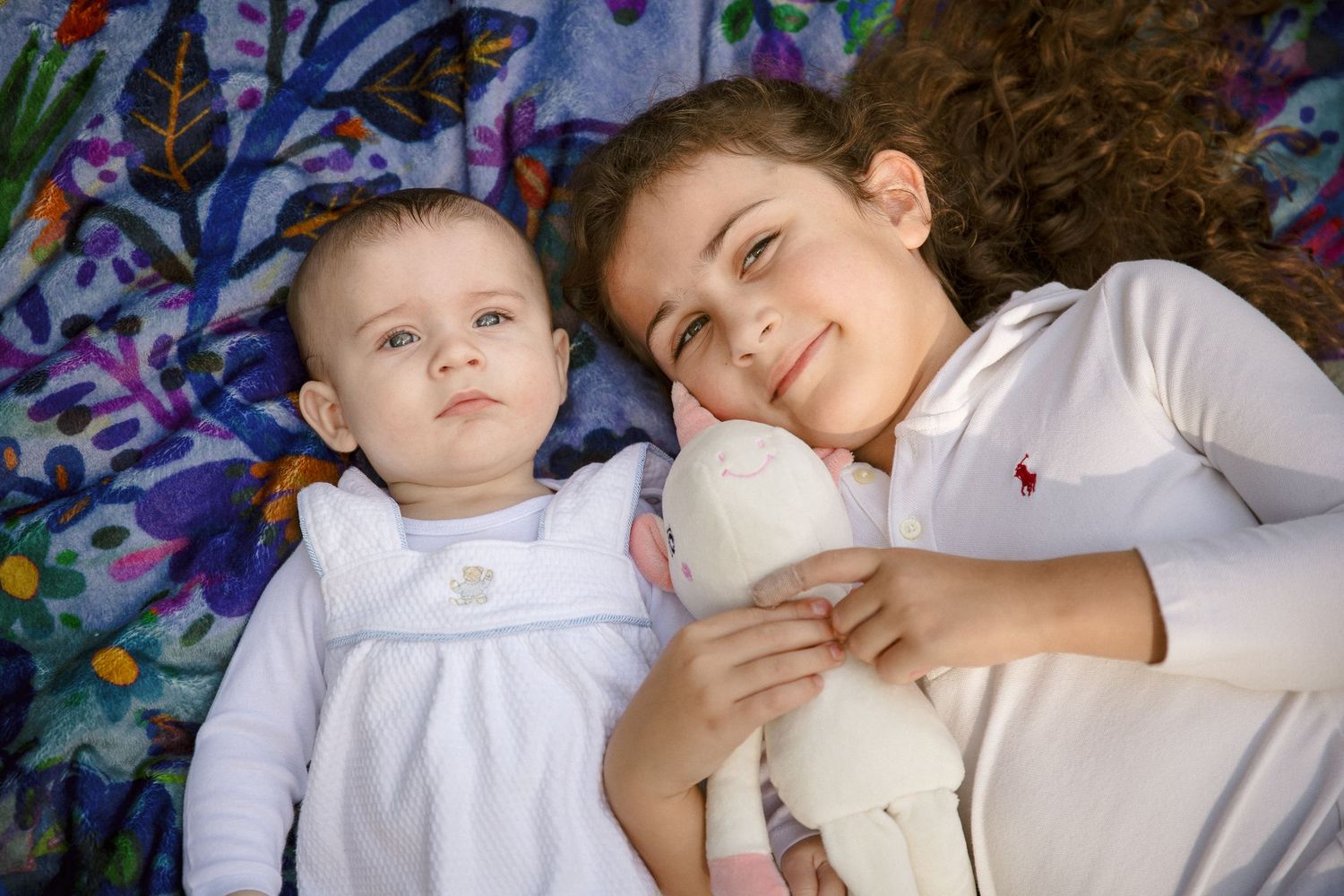 Two young sisters lying on a colorful blanket, older girl smiling and holding a stuffed animal beside her baby sibling.