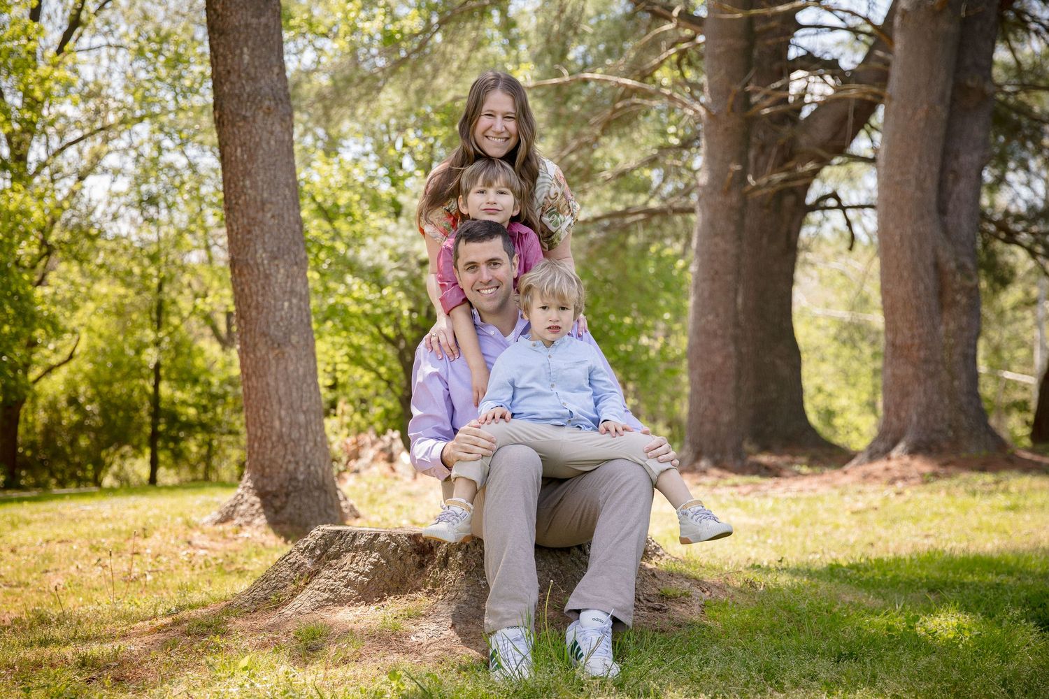 Family of four posing together on a tree stump in a lush green park, smiling for a portrait photo.