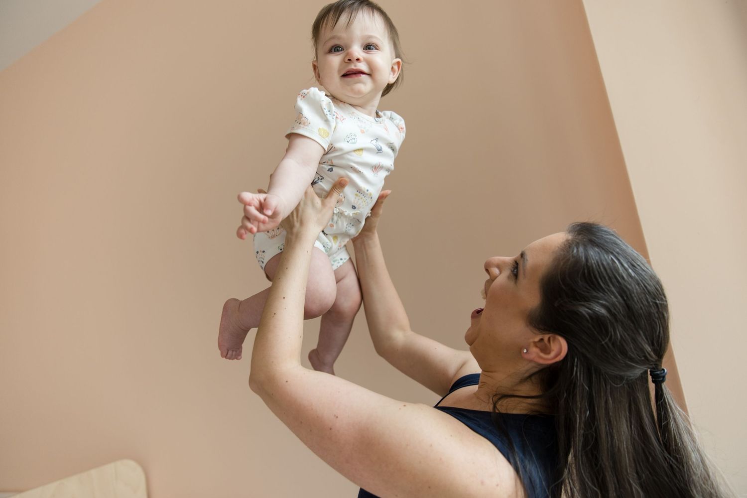 A joyful mother lifts her smiling baby overhead in a warmly lit room, sharing a playful bonding moment.