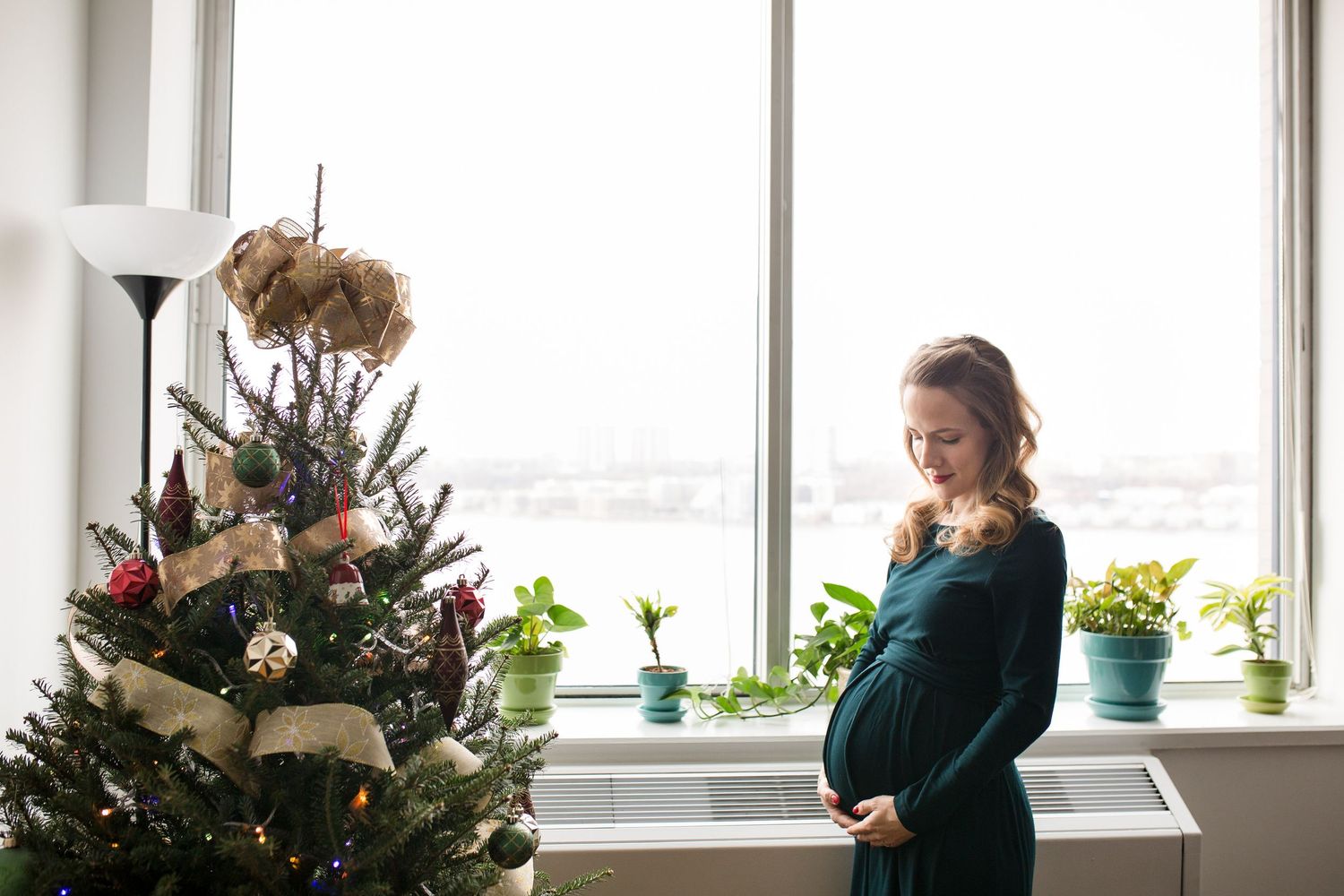 Pregnant woman in green dress holding her belly beside a decorated Christmas tree near a bright window.