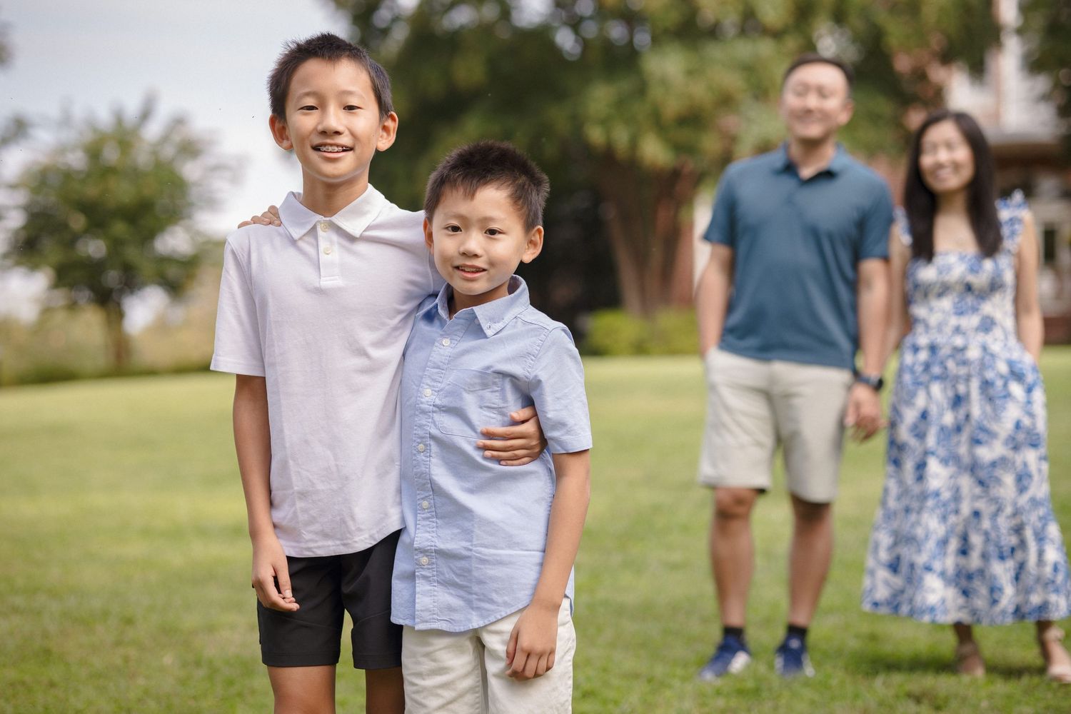 Two Asian boys smile outdoors while their parents stand behind them on a green lawn.