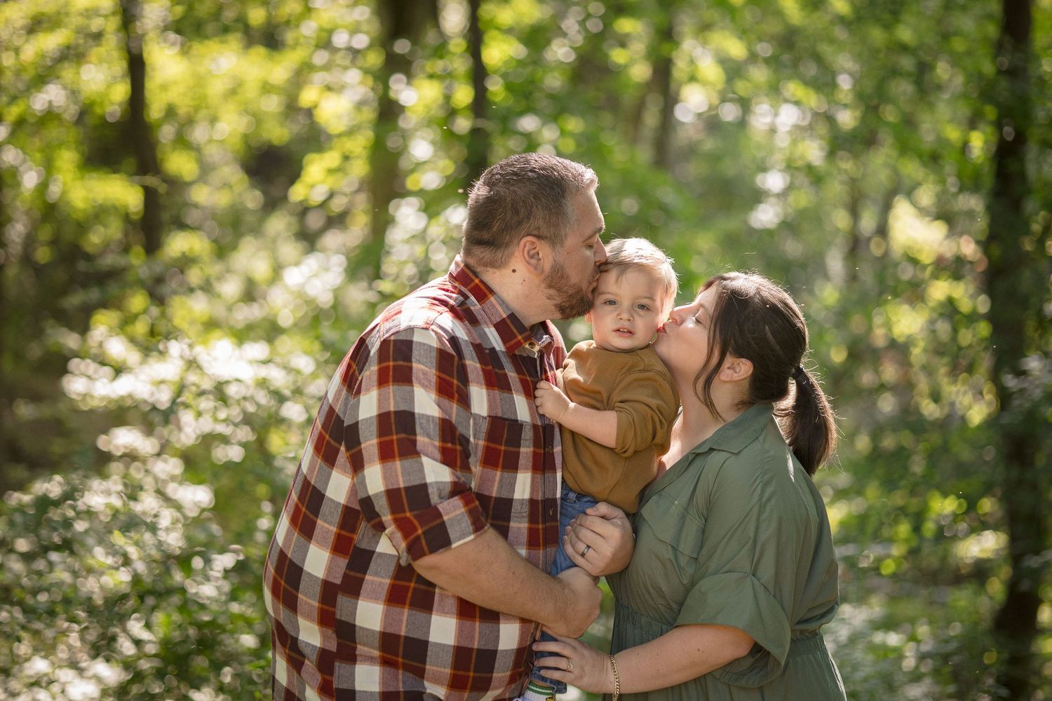 A family of three poses in a sunlit forest, parents kissing their toddler on the cheeks outdoors.